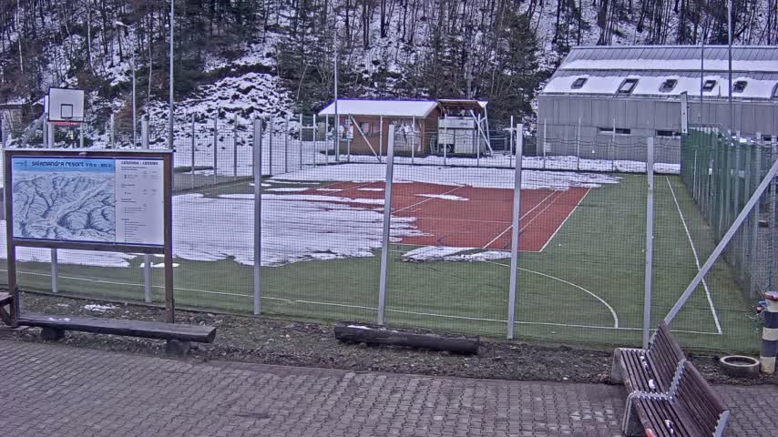 Under wintry conditions, an outdoor multi-sport court with green turf and a red tennis area is partially covered in snow and surrounded by a fence, with snow-dusted trees and buildings visible in the background.