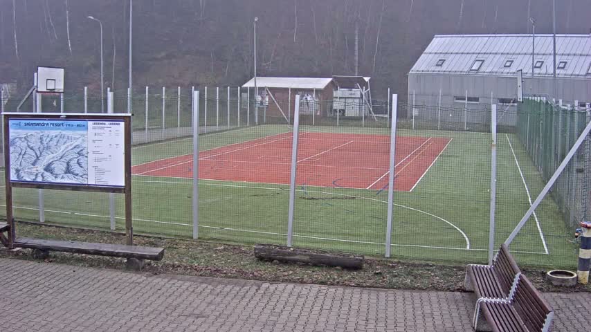 Under wintry conditions, an outdoor multi-sport court with green turf and a red tennis area is partially covered in snow and surrounded by a fence, with snow-dusted trees and buildings visible in the background.