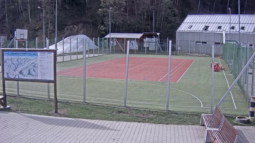 Under wintry conditions, an outdoor multi-sport court with green turf and a red tennis area is partially covered in snow and surrounded by a fence, with snow-dusted trees and buildings visible in the background.