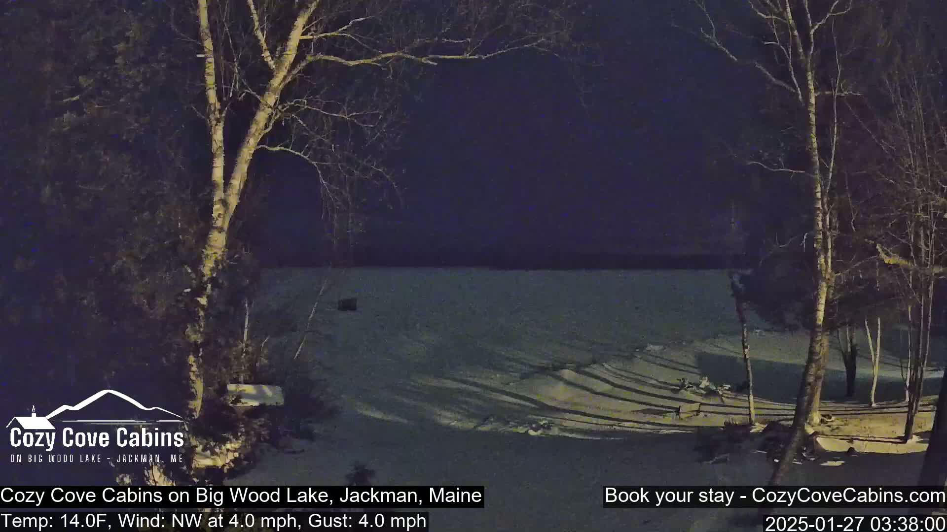 A nighttime view of a snow-covered lakeshore, with bare trees silhouetted against a dark sky, under a cold 14°F temperature with a northwest wind.