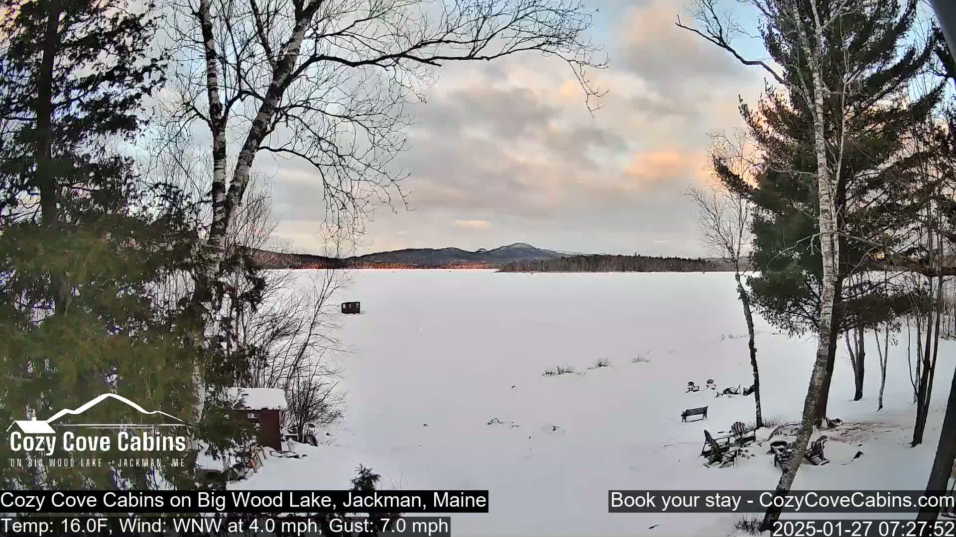 A snow-covered lake is seen from a snowy shoreline under a partly cloudy sky with a hint of sunset.