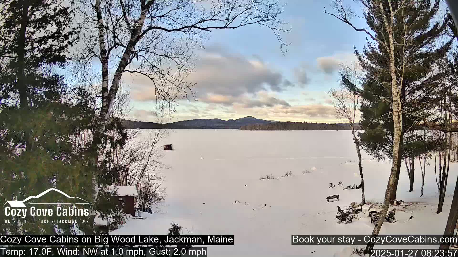 A snow-covered frozen lake is seen from a lakeside vantage point under a partly cloudy sky, with bare trees and a small cabin visible in the foreground.
