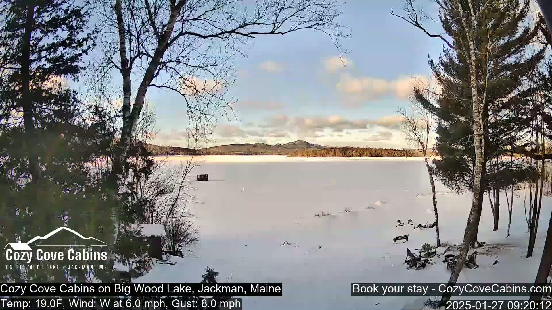 A snow-covered frozen lake is seen from a lakeside cabin under a partly cloudy sky with a light wind.