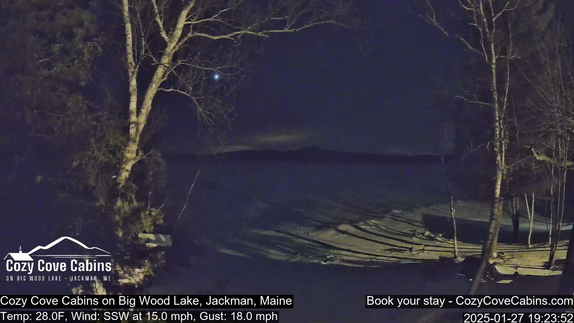 A nighttime view of a snow-covered lakeshore with bare trees under a dark sky, with a 15 mph wind from the south-southwest and a temperature of 28°F.