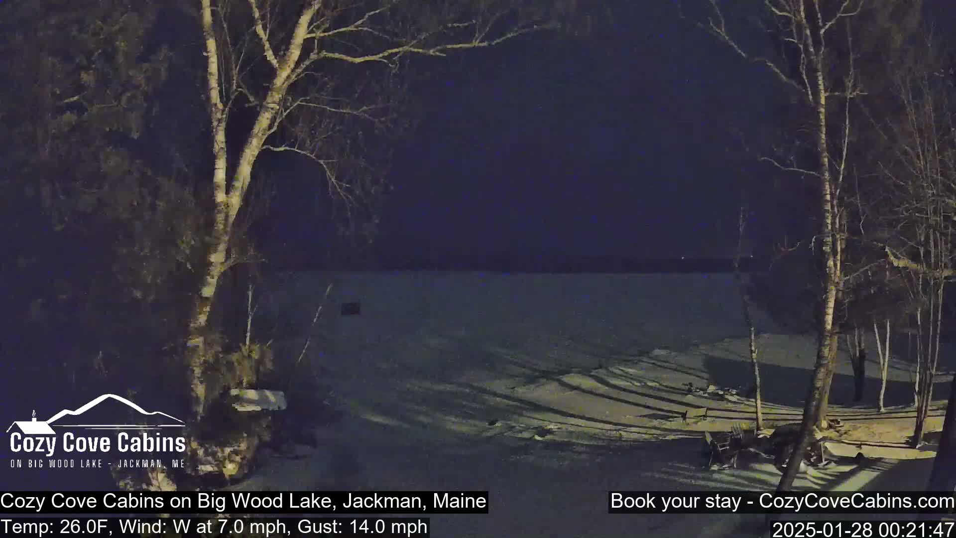 A nighttime view of a snow-covered lake, partially frozen, with bare trees silhouetted against a dark sky under a windy, 26-degree Fahrenheit conditions.