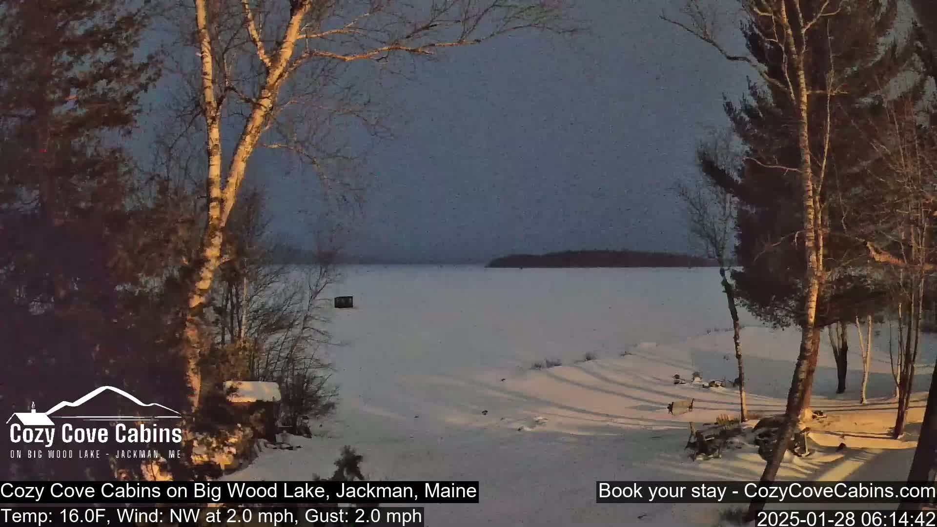 A snow-covered landscape shows a frozen lake at dusk, with bare trees in the foreground and background under a twilight sky, a light northwest wind blowing.