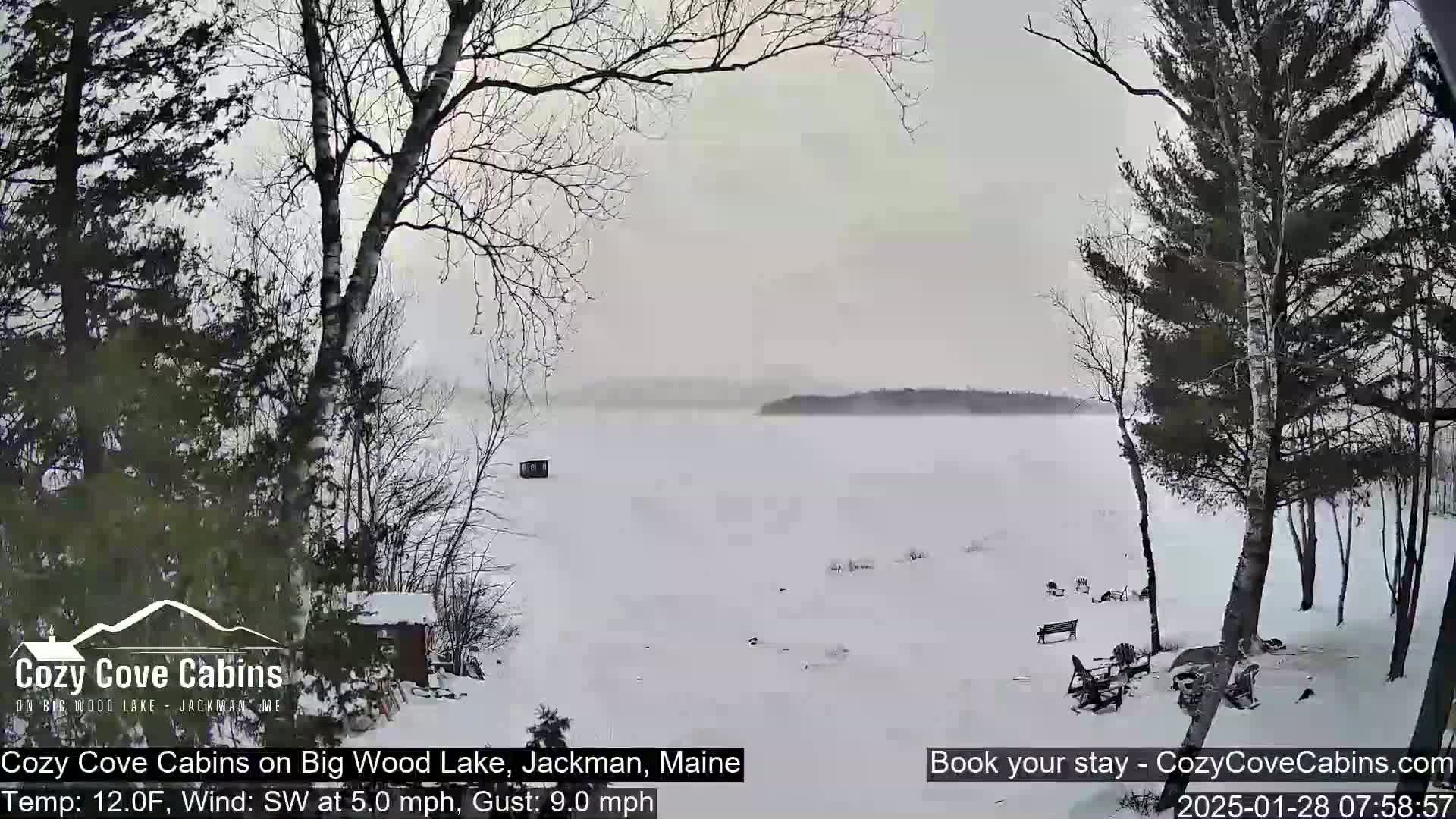 A snow-covered lake is seen from a snowy shoreline with leafless trees, a small structure on the lake's surface, and some outdoor furniture in the foreground.