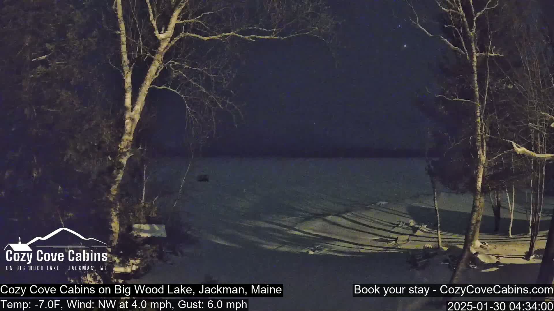 A snow-covered lakeshore at night, viewed from between two bare trees, under a dark, clear sky with a few stars visible, with a northwest wind at 4 mph and gusts up to 6 mph, and a temperature of -7°F.