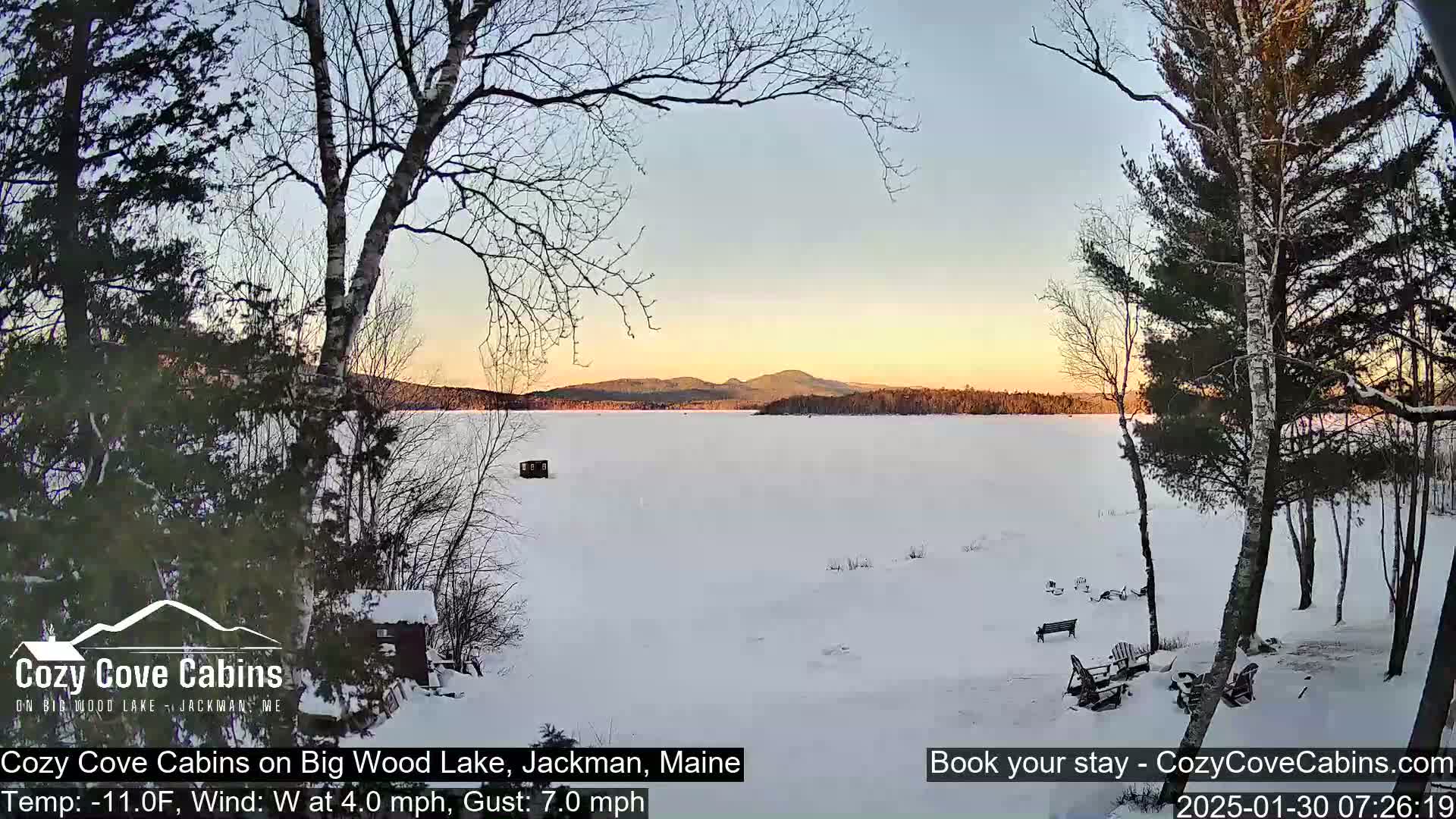 A snow-covered frozen lake at sunset, viewed from a snowy wooded shoreline with bare trees.