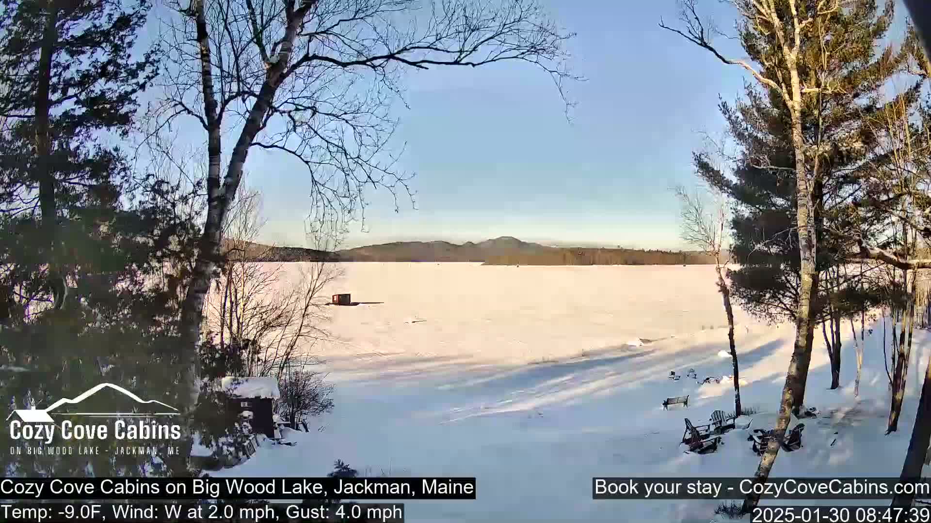 A snow-covered frozen lake is seen from a snowy shoreline, with bare trees and a sunny, clear sky in the background.