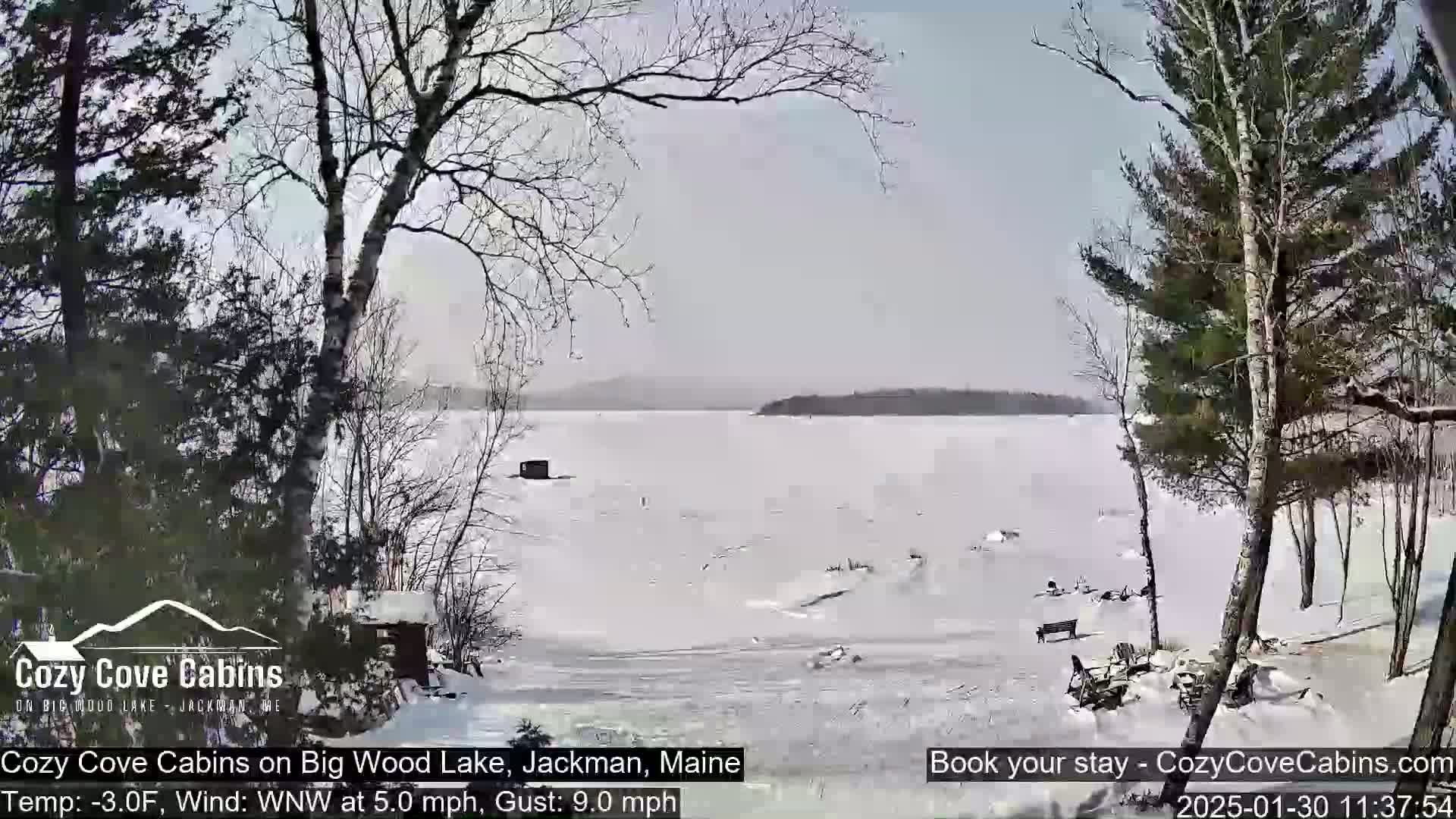 A partially frozen lake is seen from a snowy shoreline, with bare trees and a cabin visible in the foreground under a mostly clear sky.