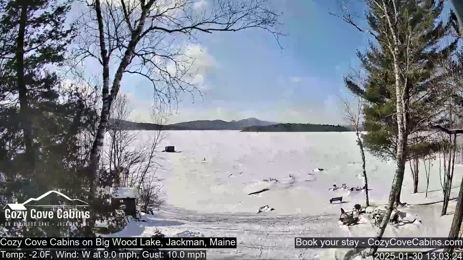 A snow-covered lake stretches to distant hills under a sunny sky, with leafless trees lining a snowy shoreline.