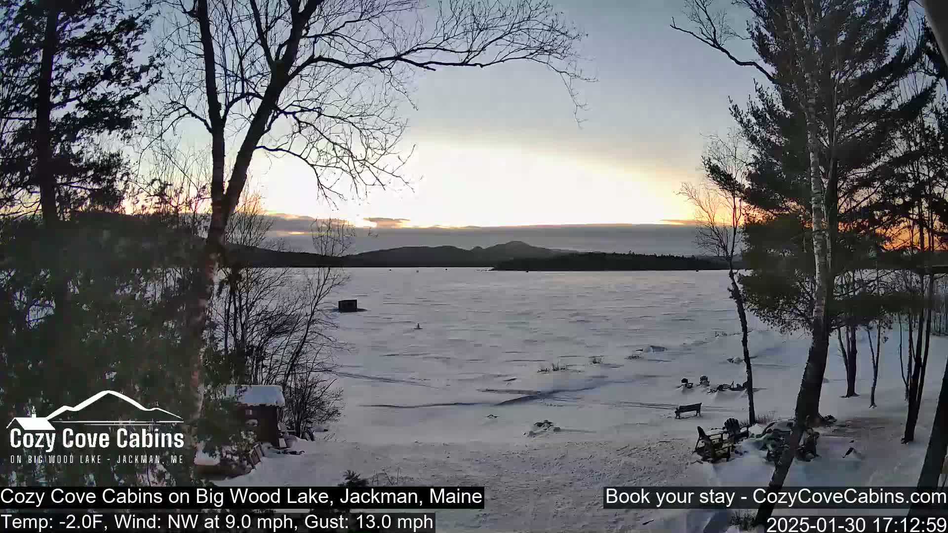 A partially frozen lake at sunset is viewed from a snowy shoreline with bare trees in the foreground under a mostly clear sky.