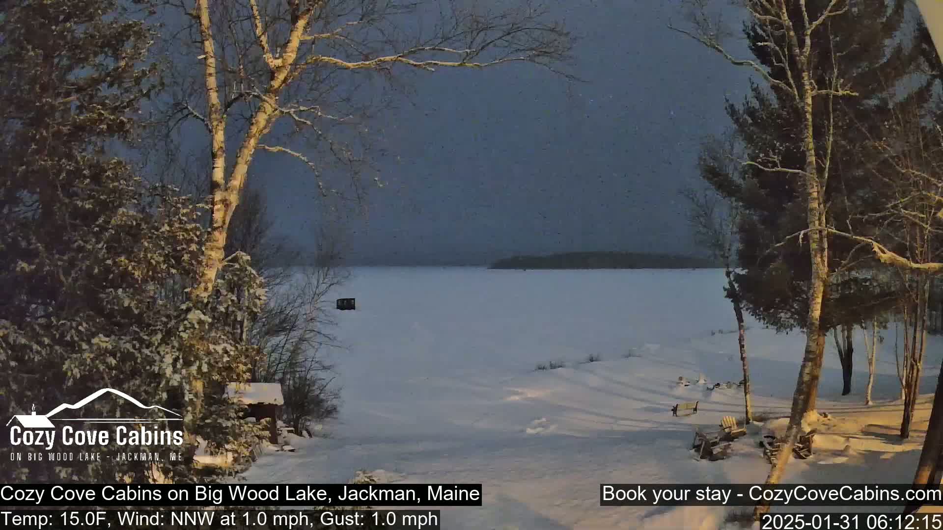 A snow-covered landscape shows a frozen lake under a twilight sky with light snow falling.