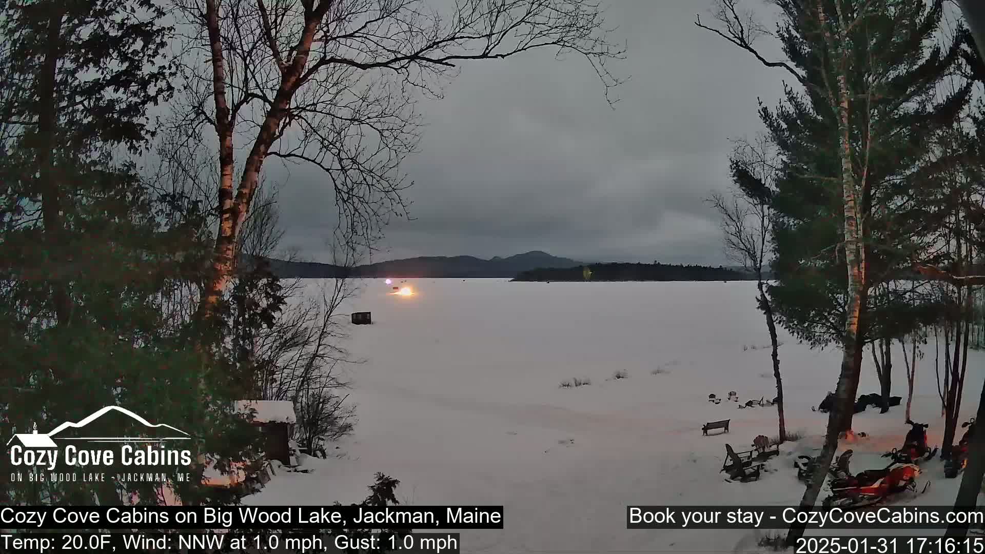 A snow-covered lake is viewed from a snowy shoreline, under an overcast sky, with bare trees and snowmobiles visible in the foreground.