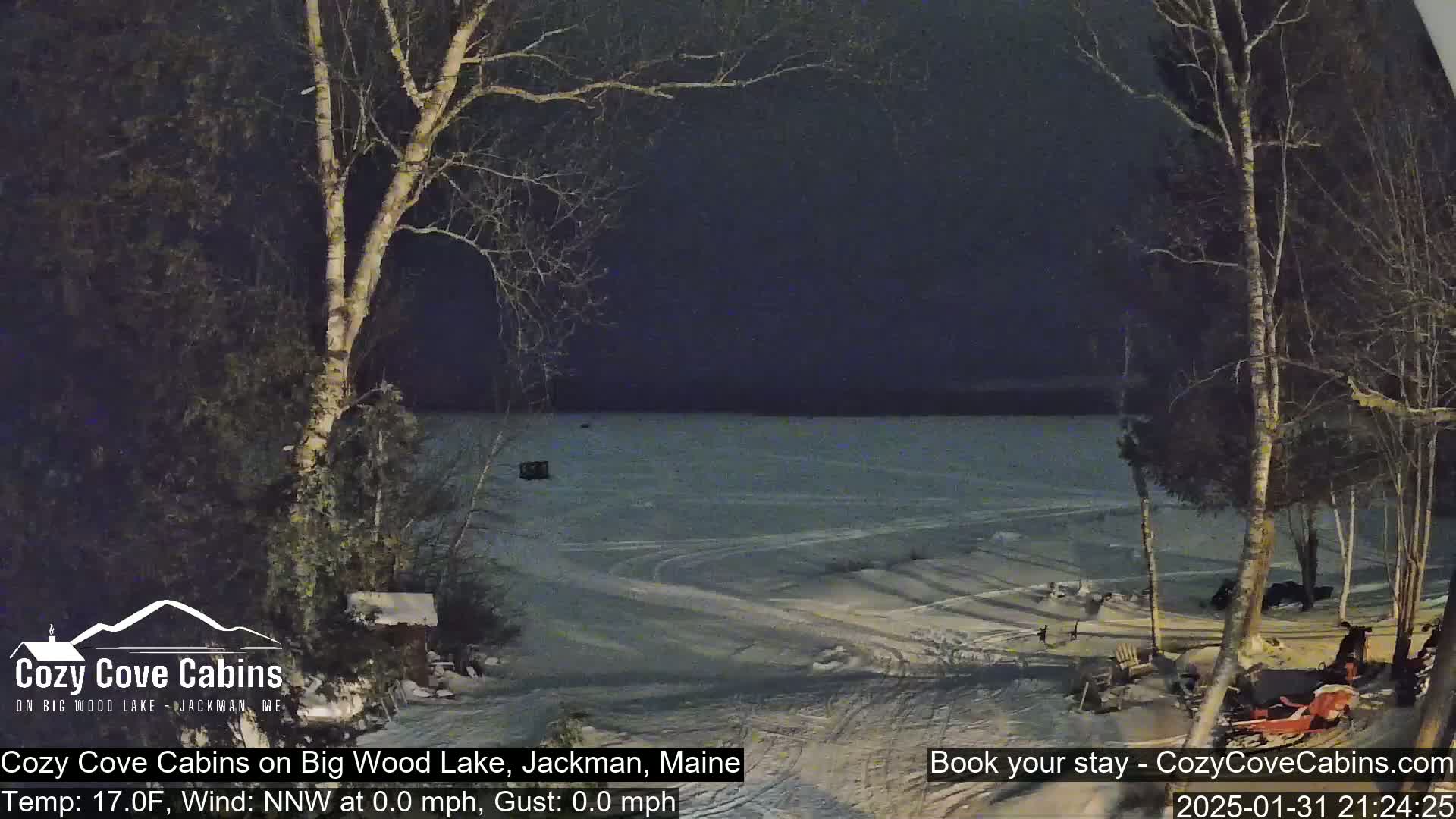 A snow-covered lake is visible at night, with bare trees and a snowmobile in the foreground under a dark sky.