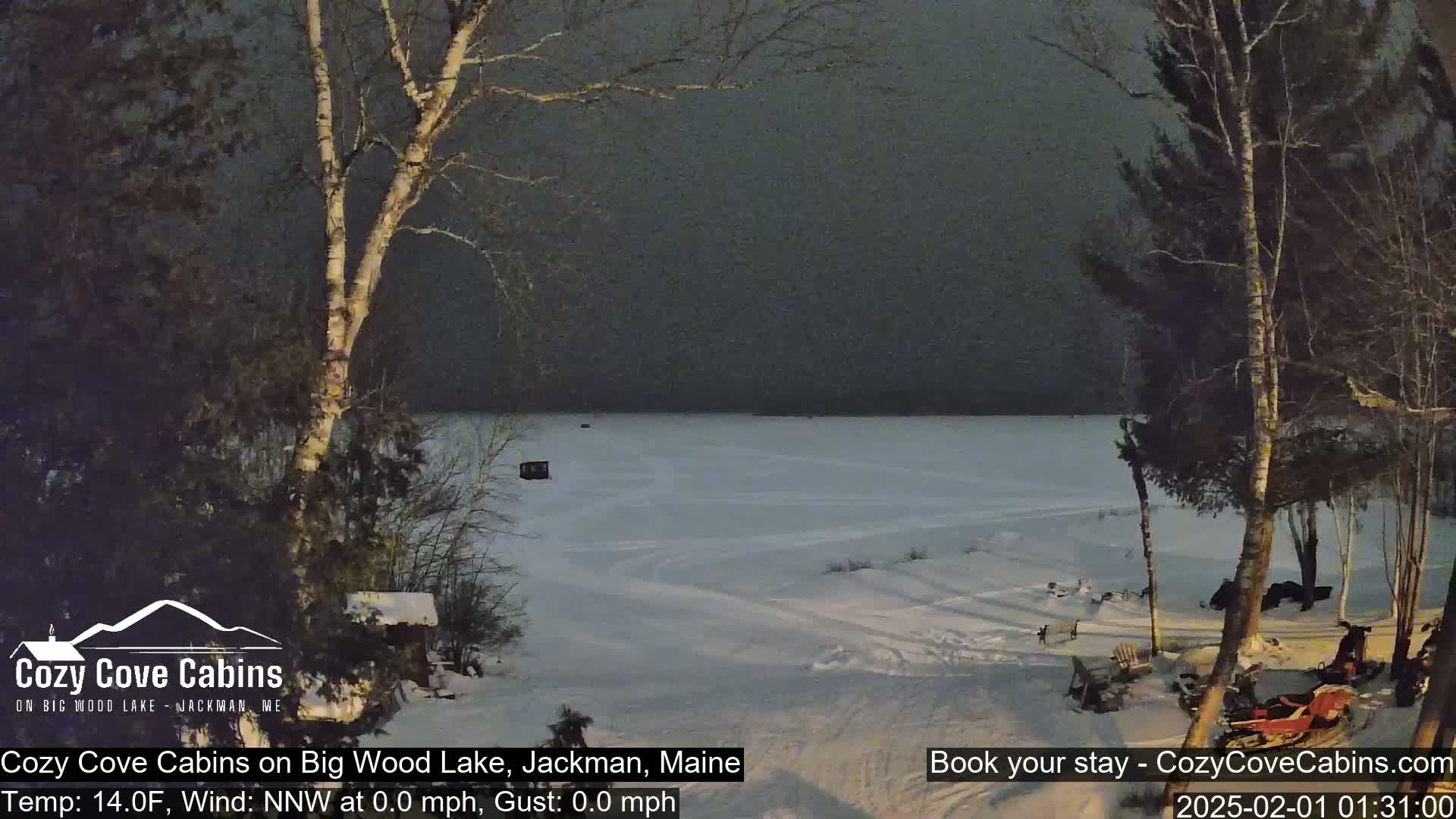 A snow-covered frozen lake is seen at night under a dark, overcast sky, with trees and snowmobiles visible in the foreground.