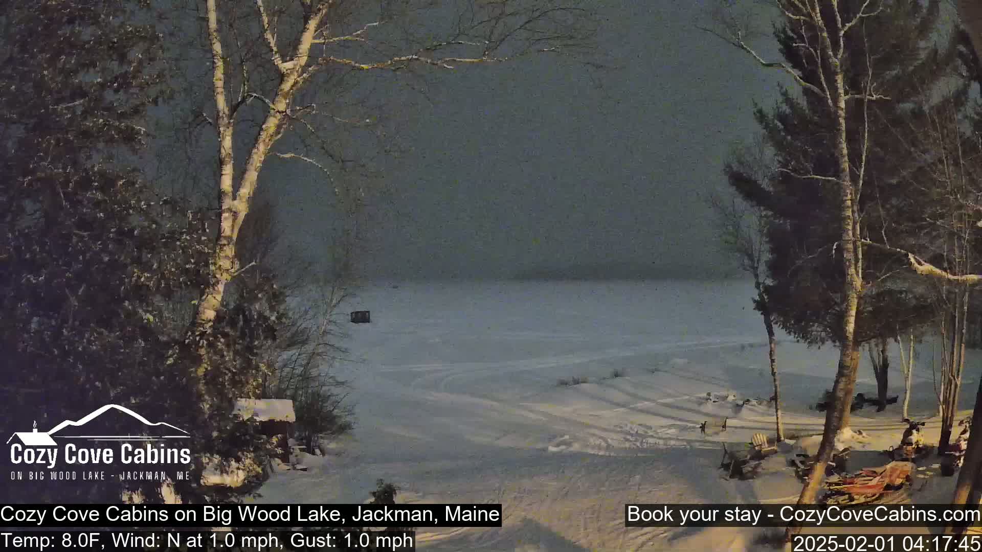 A snow-covered landscape at night, showing a frozen lake, trees covered in snow, and a cabin, under a dark, cloudy sky.