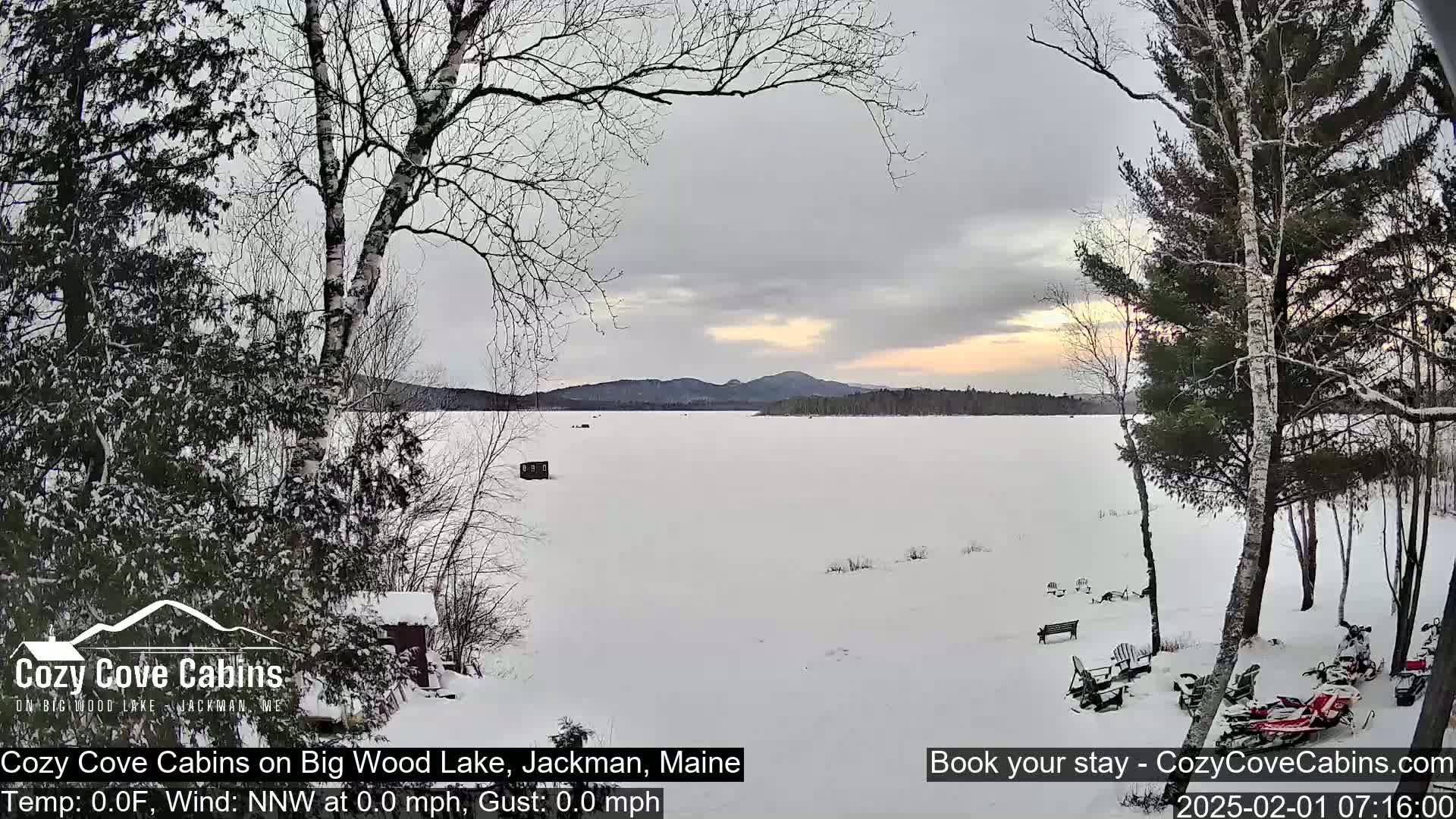 A snow-covered frozen lake is seen from a snowy lakeside vantage point, with calm weather and a pale sunrise illuminating the distant mountains.