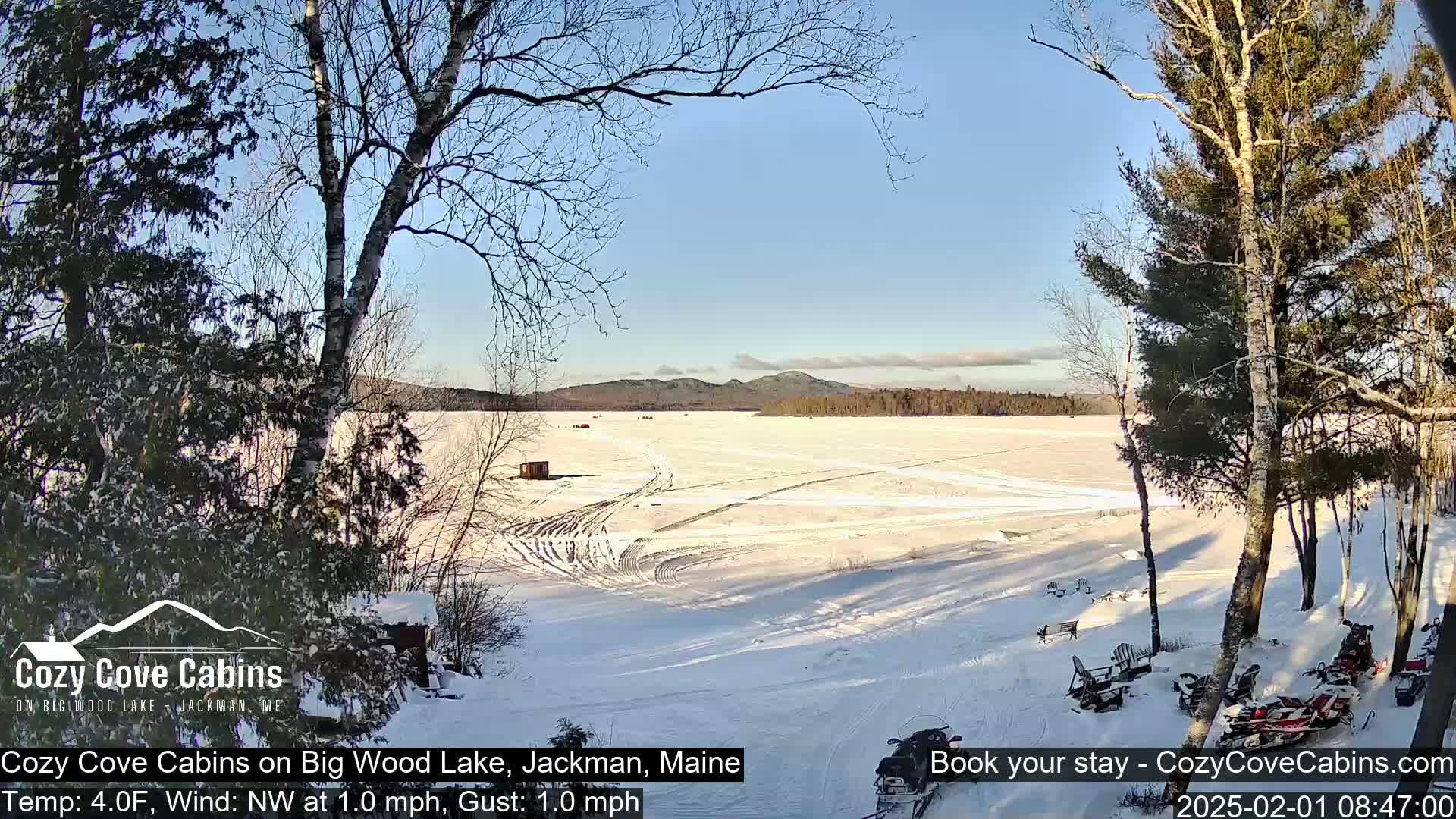 A snow-covered frozen lake is seen from a snowy shoreline, with trees, a small building, and snowmobiles visible under a clear blue sky.