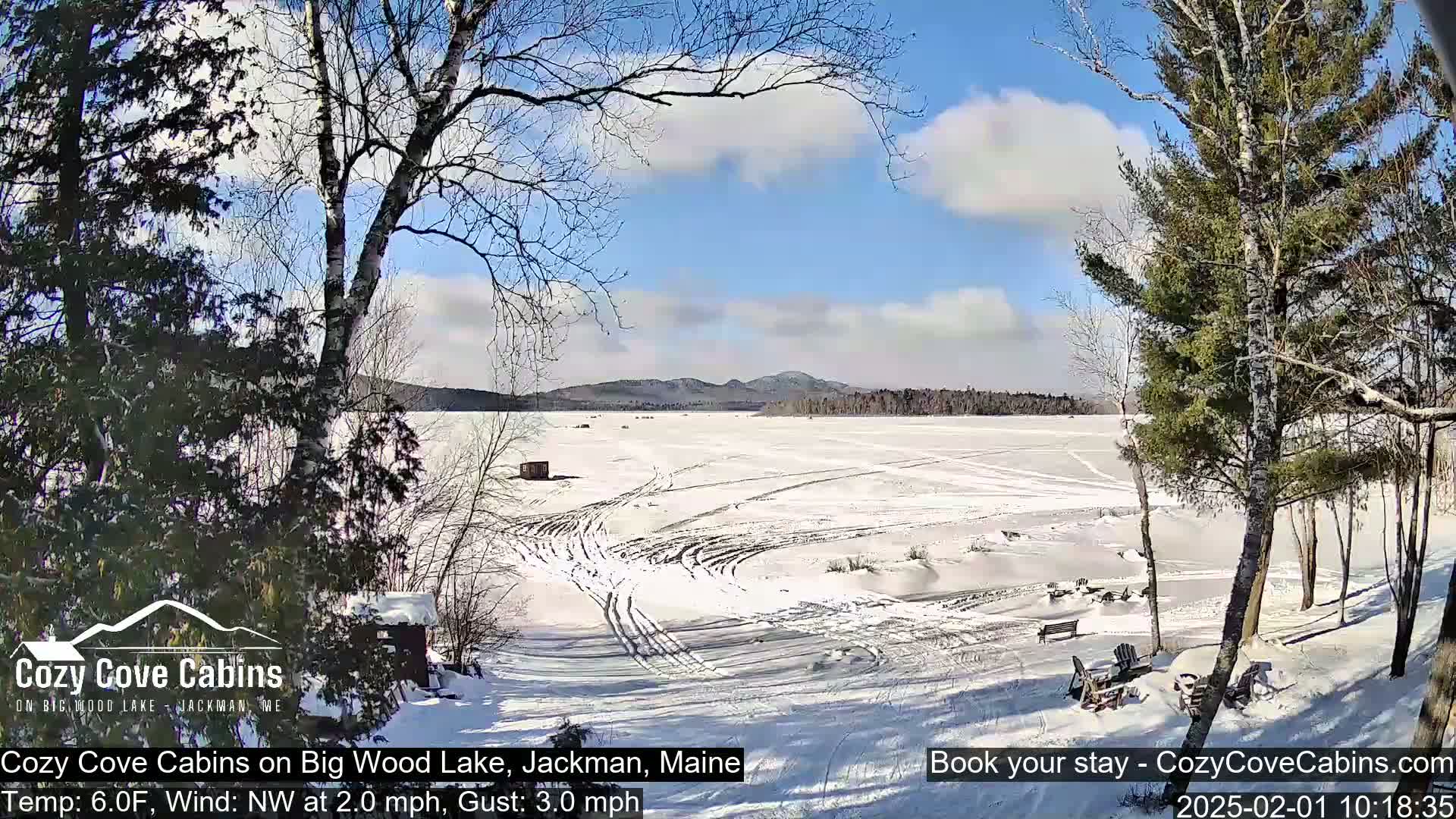 A snow-covered frozen lake is seen from a snowy lakeside vantage point under a partly cloudy blue sky.