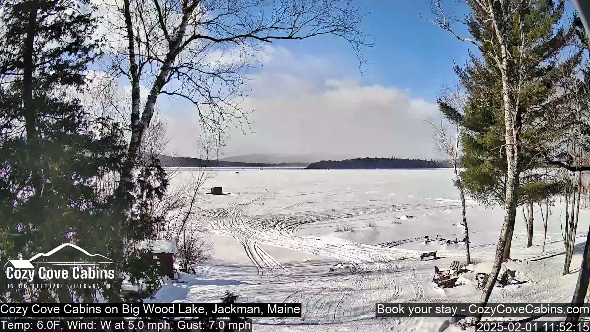 A partly cloudy, sunny winter day shows a frozen lake with snow-covered shorelines and tire tracks leading to a lakeside cabin; several trees are visible.