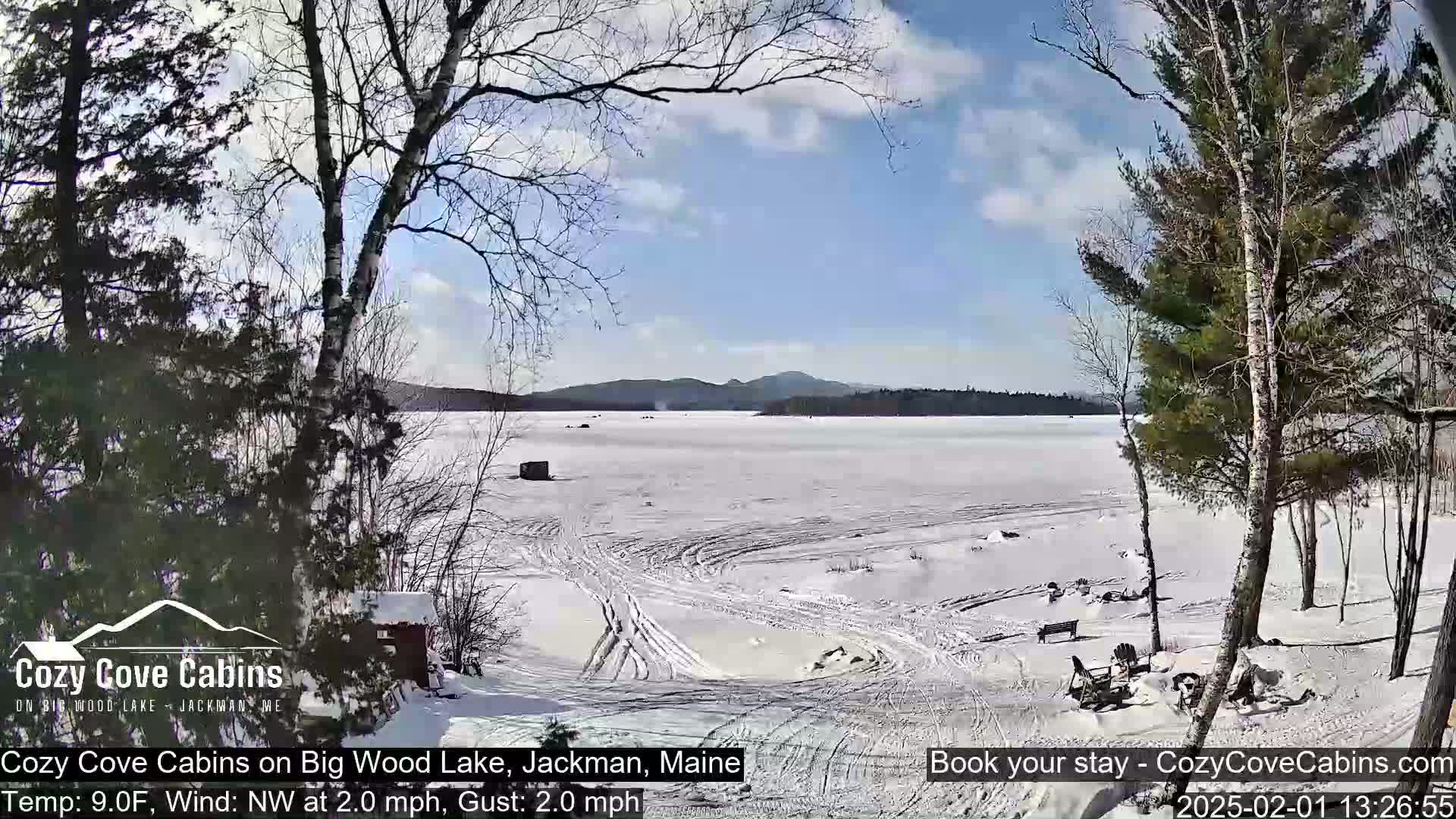 A snow-covered frozen lake is seen from a snowy shoreline under a partly cloudy, sunny sky.