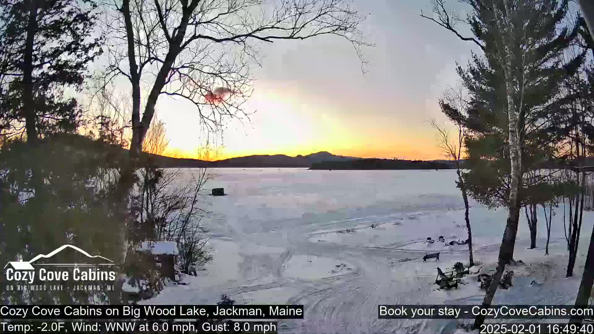 A frozen lake at sunset, partially covered in snow, is viewed from a snowy shoreline with bare trees and a cabin in the foreground; the sky is a mix of orange, yellow, and purple.