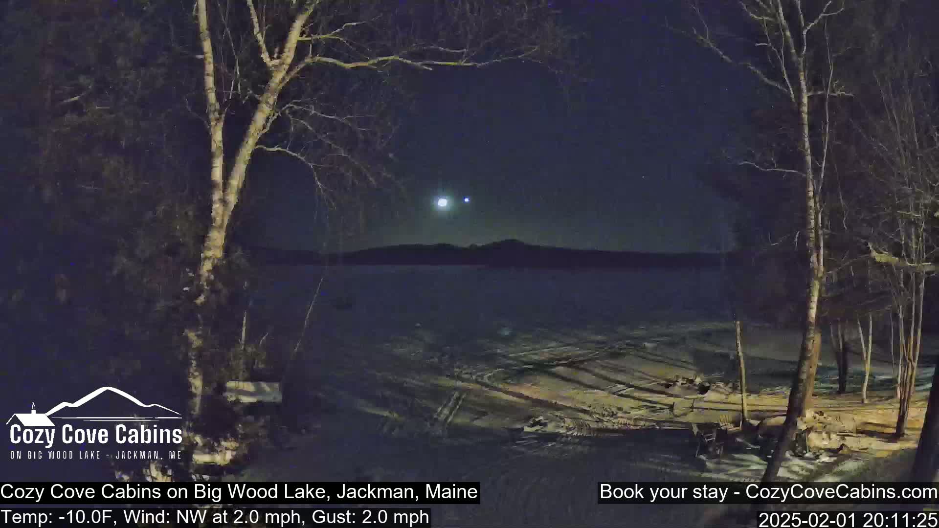 A nighttime view of a snow-covered lake under a clear, dark sky with a few bright stars and a distant mountain range, framed by bare trees in the foreground.