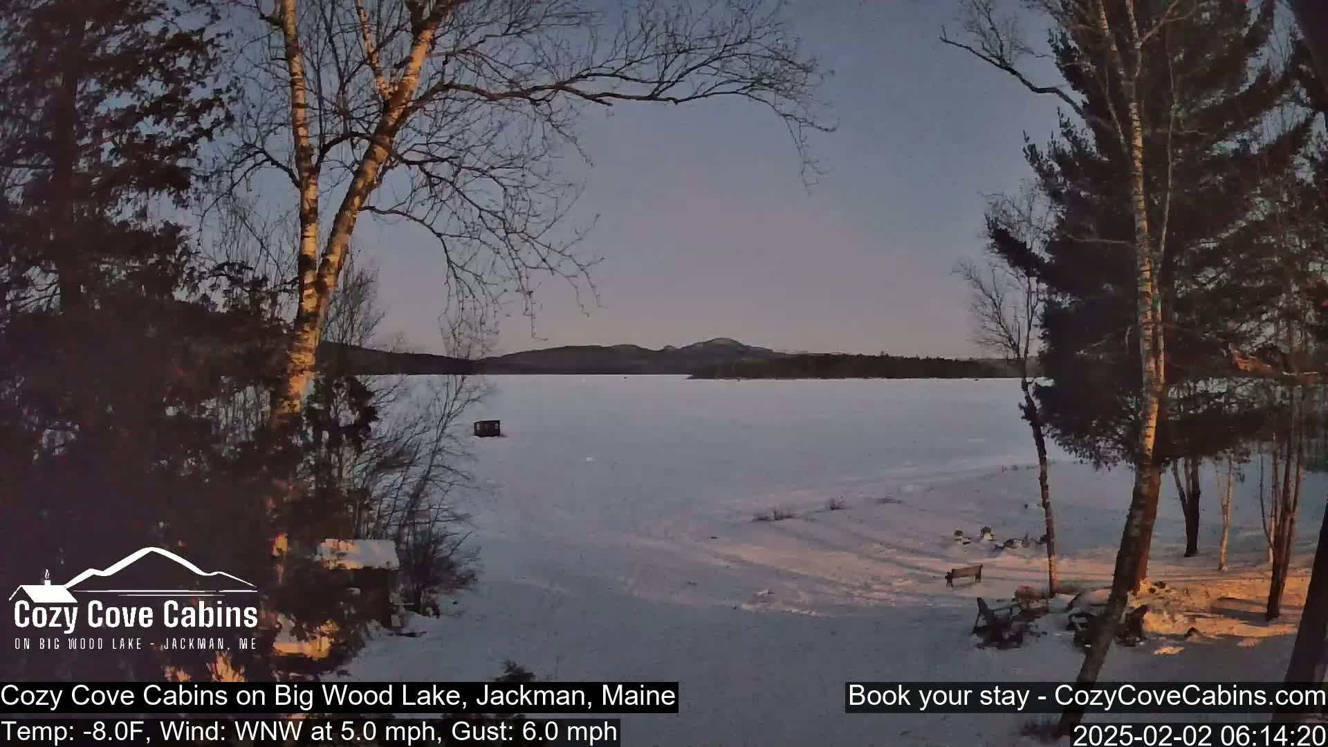 A frozen lake is seen at twilight from a snowy shoreline, with bare trees and a few structures visible.