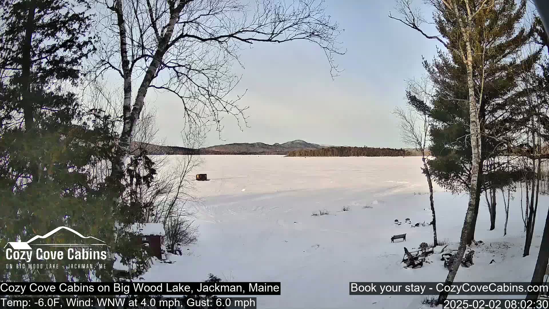 A snow-covered frozen lake is seen from a snowy lakeside vantage point under a mostly clear sky.