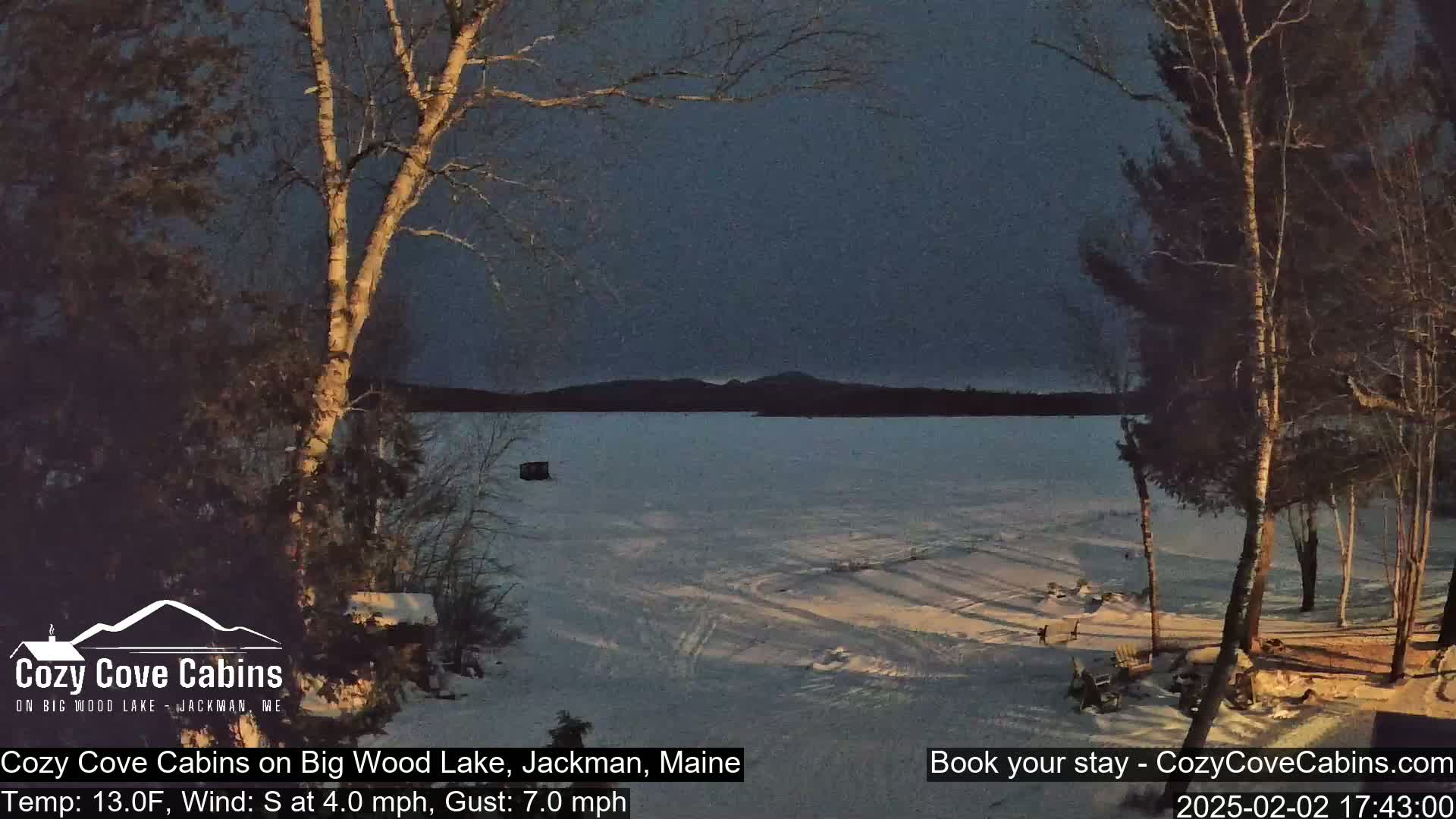 A snow-covered lake is seen at dusk under a dark sky, with bare trees in the foreground and a distant mountain range visible.