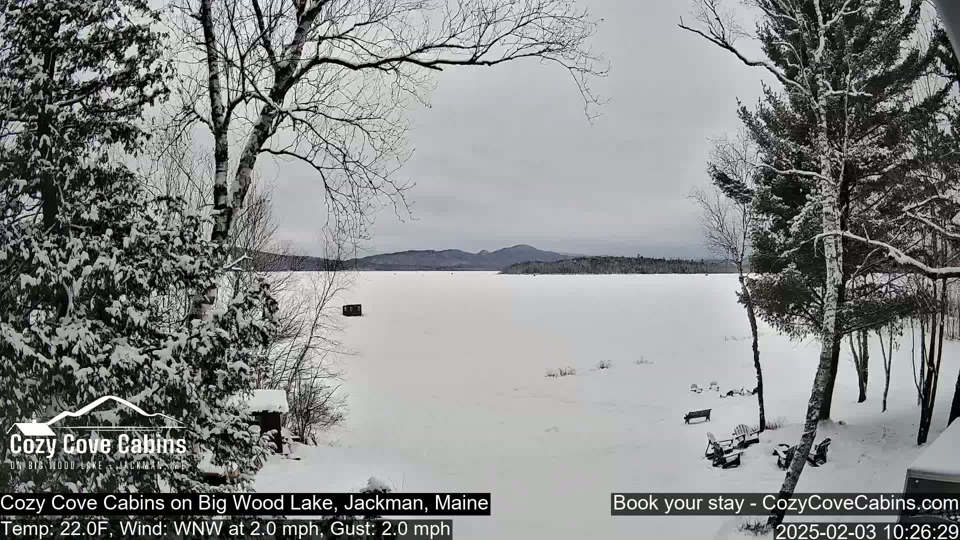 A snow-covered frozen lake is viewed from a snowy lakeside, with snow-covered trees and a small structure visible in the distance under an overcast sky.