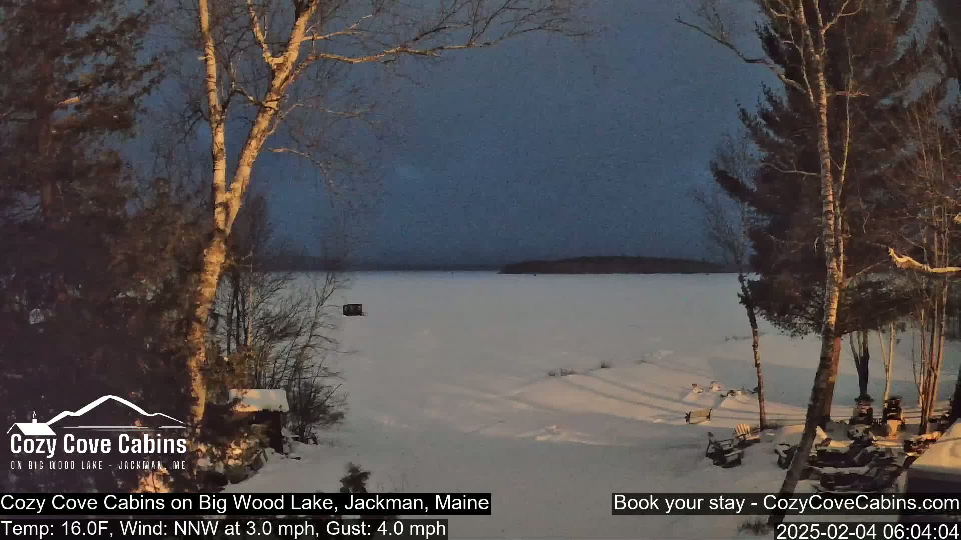 A snow-covered frozen lake is seen at twilight, with bare trees and a cabin in the foreground under a dark, mostly clear sky.