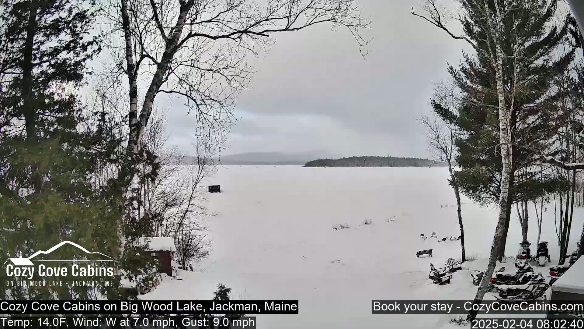 A snow-covered frozen lake is viewed from a snowy shoreline with leafless trees and snowmobiles parked nearby, under an overcast sky.