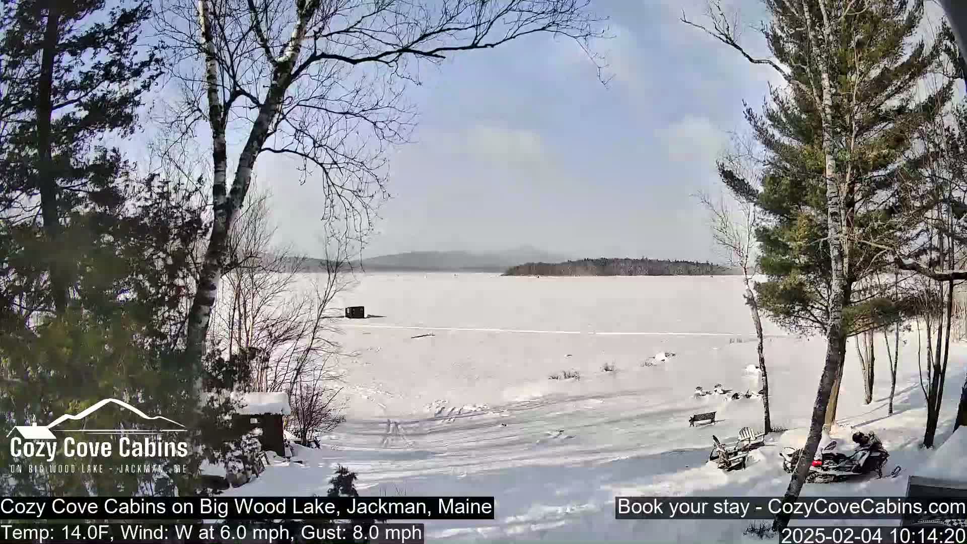 A snow-covered frozen lake is viewed from a snowy shoreline with leafless trees and a snowmobile parked nearby under a sunny sky.