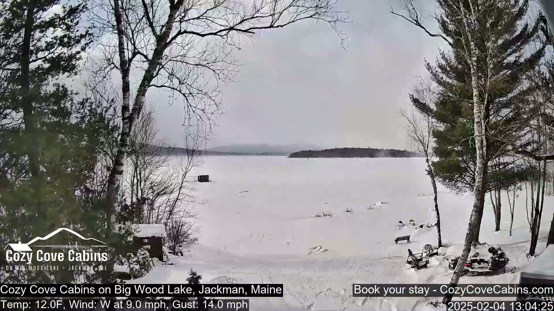 A snow-covered frozen lake is seen from a snowy shoreline with leafless trees and a snowmobile, under an overcast sky with a westerly wind.
