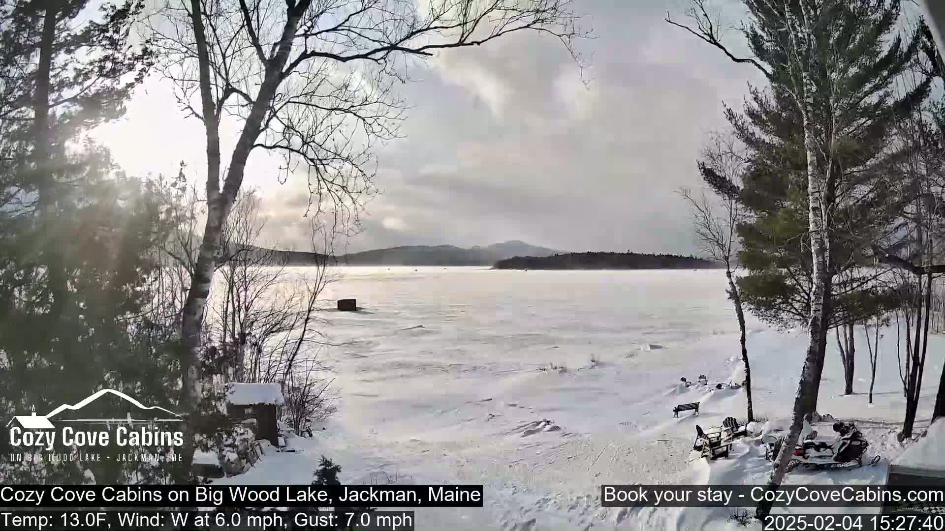 A snow-covered lake with distant hills is seen from a snowy shoreline, under a partly cloudy sky with a light wind.