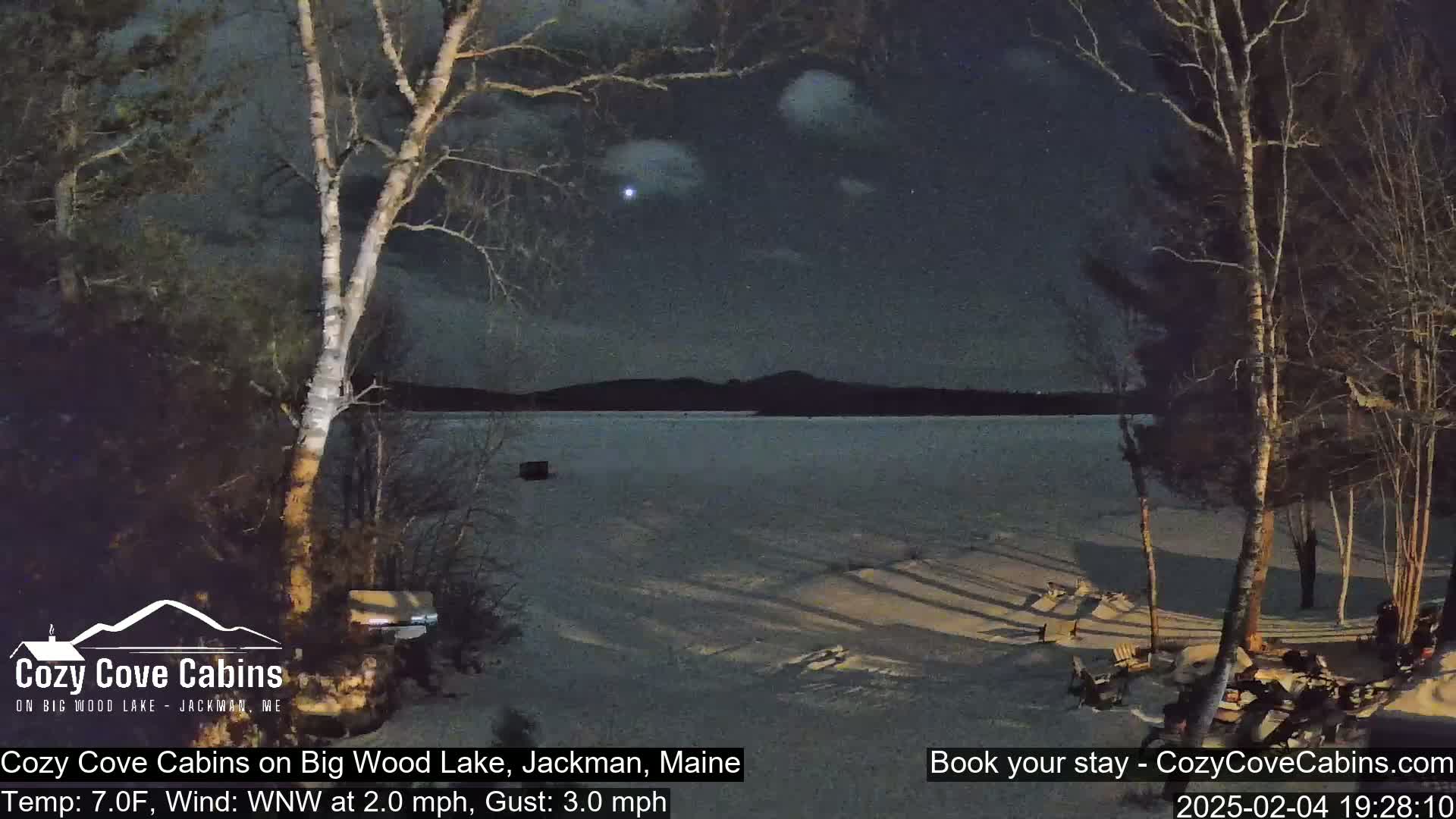 A snow-covered shoreline at night, with a partially frozen lake extending to distant hills under a partly cloudy sky, illuminated by a bright star.