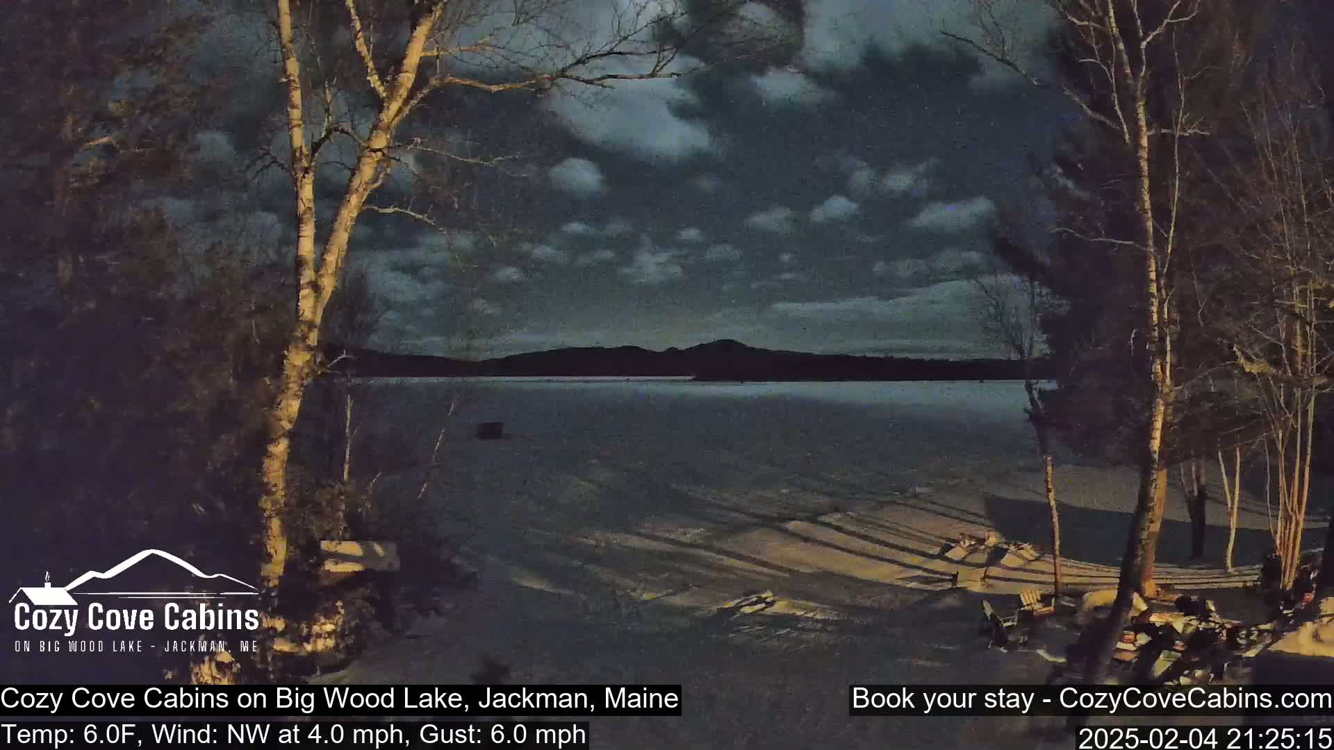 A nighttime view of a snow-covered lakeshore with leafless trees under a partly cloudy sky, with a calm lake and distant hills visible.