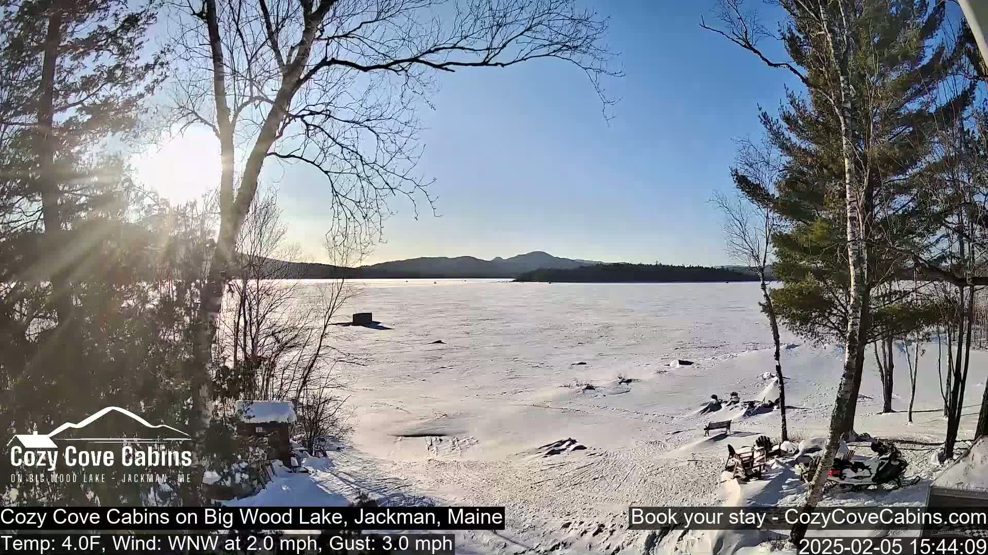 A frozen lake stretches to snow-covered mountains under a sunny, clear sky, viewed from a snow-covered shoreline with trees and a snowmobile.