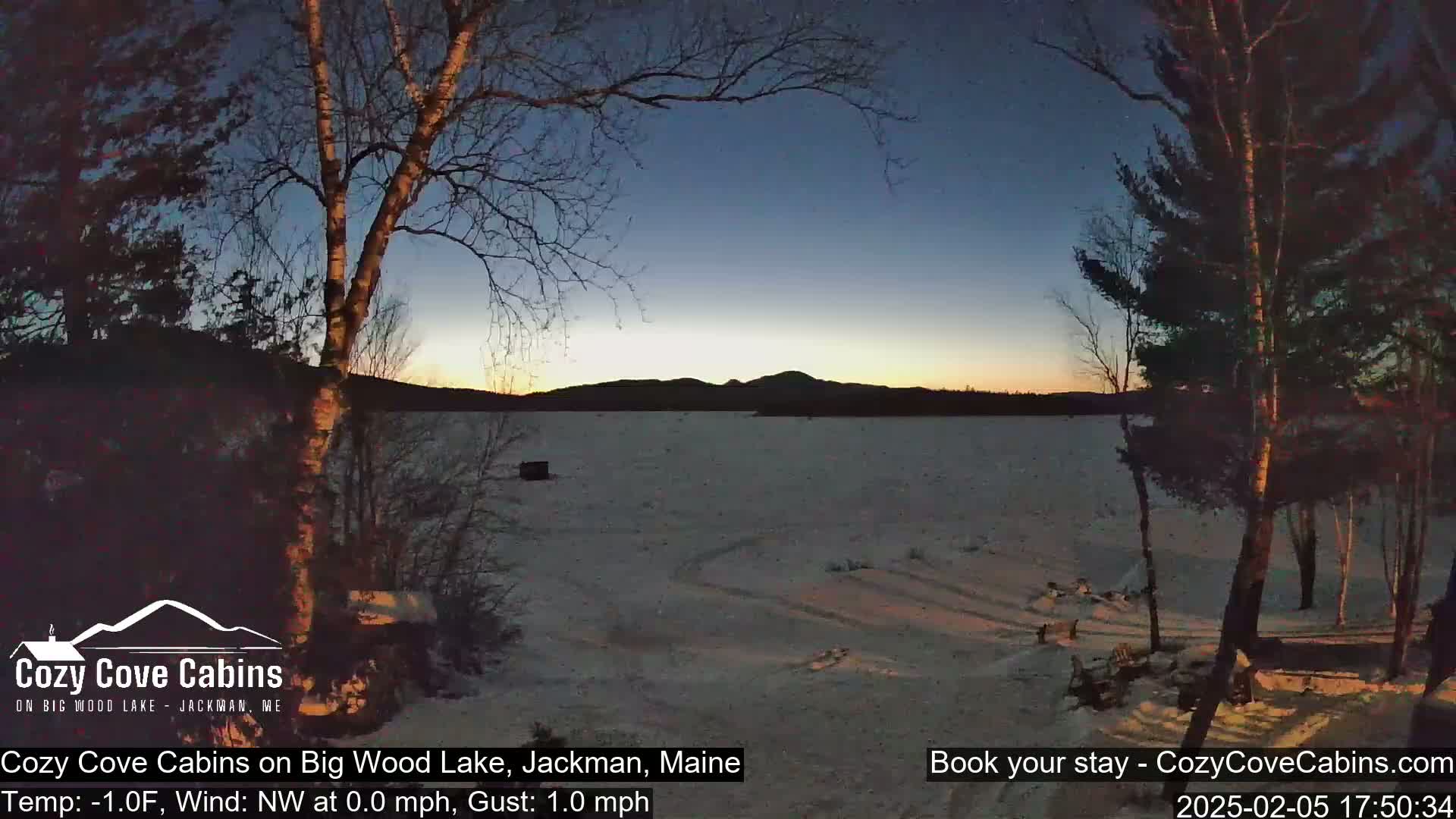 A snow-covered frozen lake at sunset, viewed from a lakeside location with bare trees silhouetted against the twilight sky.