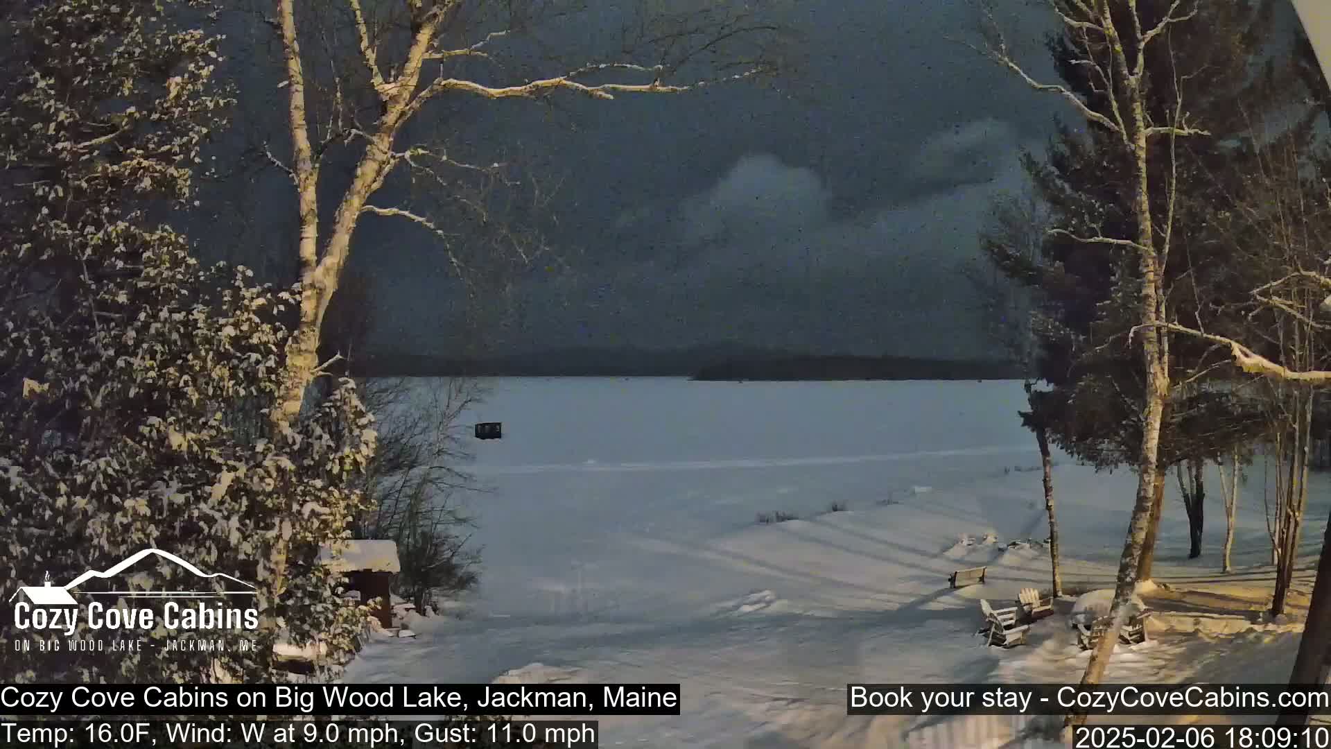 A snow-covered landscape shows a frozen lake under a dark, cloudy sky, with snow-laden trees in the foreground and a small structure visible on the lake's surface.