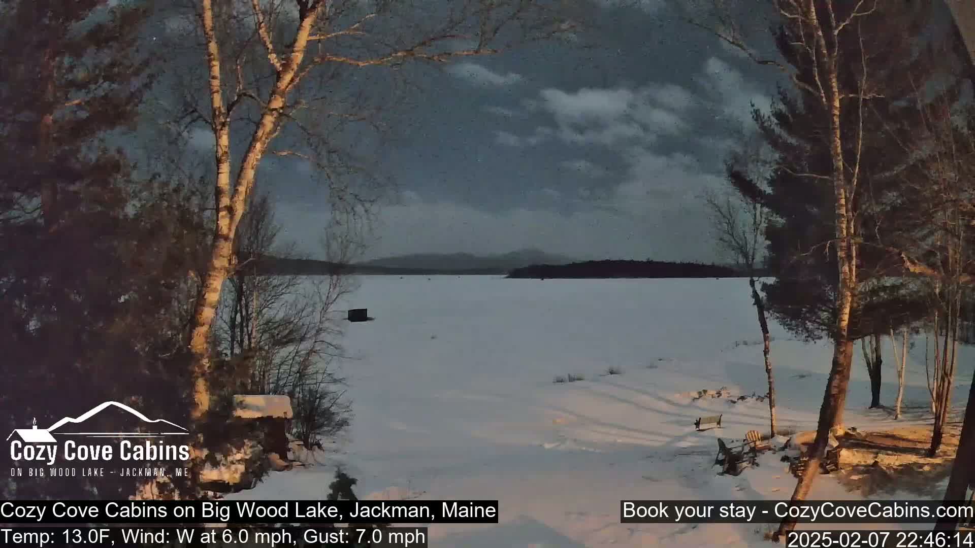 A snow-covered lake is seen at dusk under a partly cloudy sky, with bare trees in the foreground and mountains in the distance.