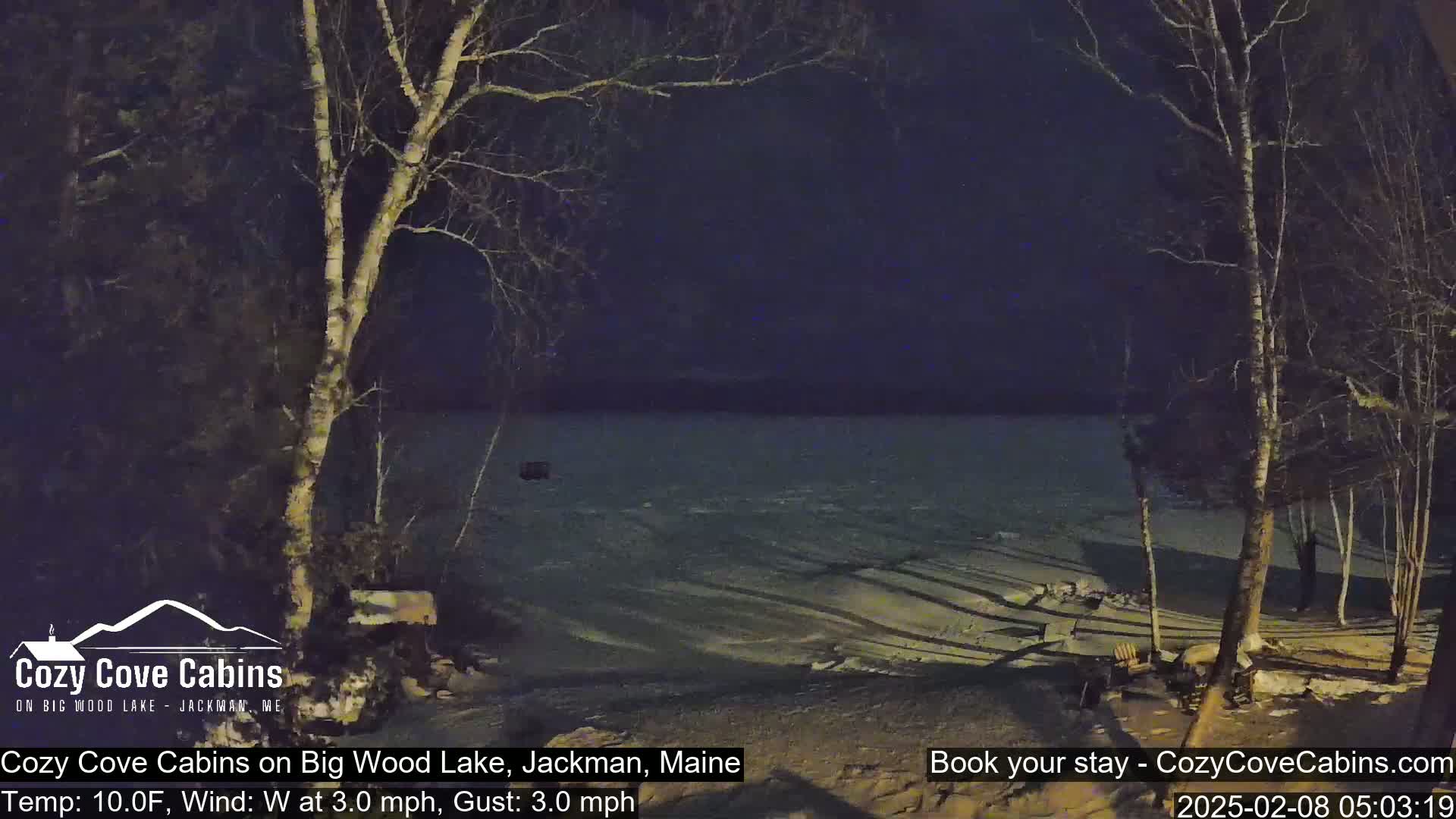 A nighttime view of a partially frozen lake, seen from a snow-covered shoreline with bare trees under a dark sky and a 10°F temperature, with a westerly wind of 3 mph.