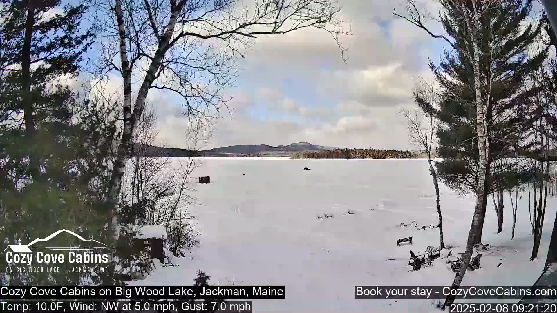 A snow-covered frozen lake is viewed from a snowy shoreline, with bare trees and a small cabin visible, under a partly cloudy sky.