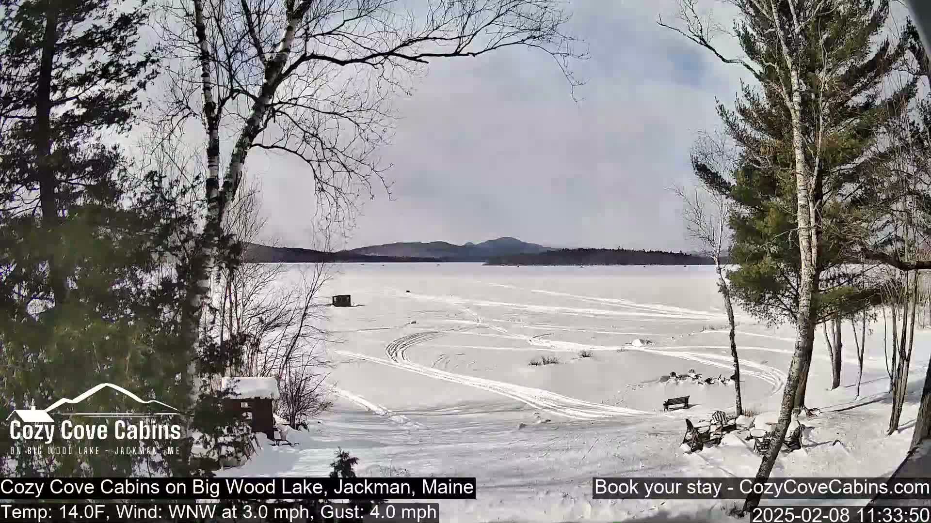A snow-covered frozen lake with trees and a few small structures is visible under a partly cloudy sky.