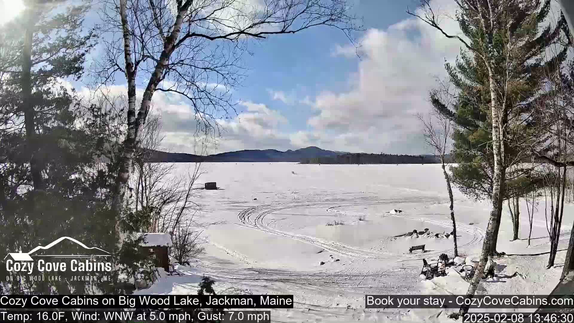 A snow-covered frozen lake is viewed from a snowy shoreline with trees and a partly sunny sky.