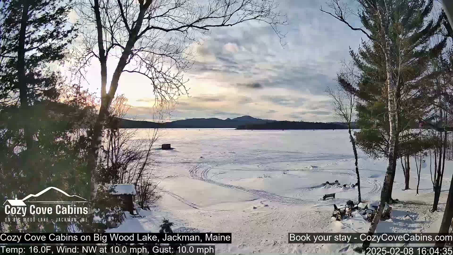 A snow-covered lake with a distant mountain range under a partly cloudy sky is seen from a snowy lakeside vantage point with leafless trees.
