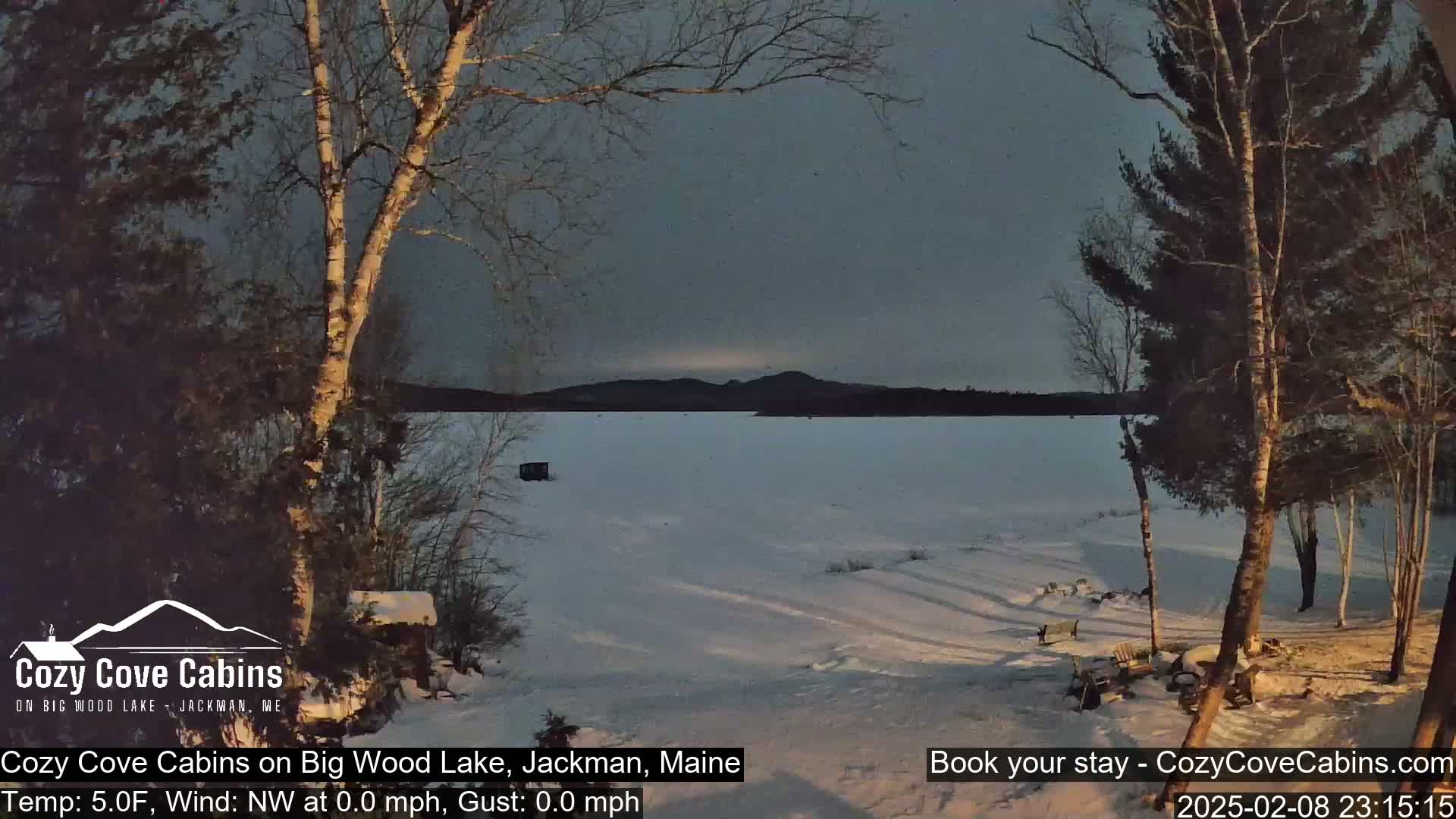 A snow-covered frozen lake is seen at dusk from a snowy shoreline, framed by leafless trees under a gray sky.